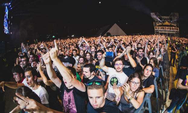 Menschenmenge auf einem Musikfestival mit erhobenen Händen, einige sitzen und andere stehen, nahe einem Metallzaun mit Gebäuden und Lichtern im Hintergrund.