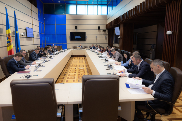 Gruppe von Menschen um einen langen Tisch versammelt mit Papieren, Büchern, Mikrofonen und anderen Gegenständen während einer Sitzung des Europäischen Parlaments in Brüssel, mit Fahnen links und Bildschirmen und Lichtern im Hintergrund.