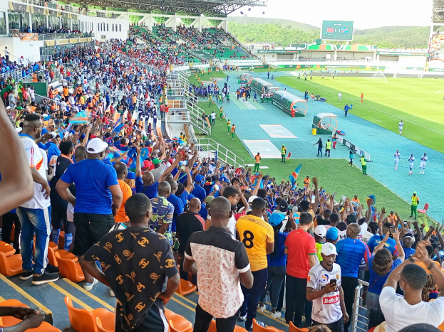 Eine große Menschenmenge versammelt sich vor einem eingezäunten Fußballfeld unter einem klaren blauen Himmel, mit Hügeln im Hintergrund, einige sitzen auf Stühlen und andere stehen, in Erwartung eines Spiels.