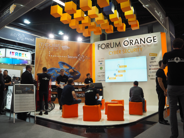 Eine Gruppe von Menschen an einem IT-Cube Systems Forum Orange Cyber Security Stand, einige sitzen auf Stühlen und andere stehen, mit einer Tafel, einem Bildschirm, Regalen mit Gegenständen, Schildern, einer Decke mit Deckenleuchten und Lampen.
