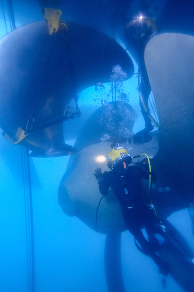 Ein Tauchender in einem Neoprenanzug, Sauerstoffflasche und Helm arbeitet an einem großen Propeller im Meer, umgeben von Seilen und Meereslebewesen.