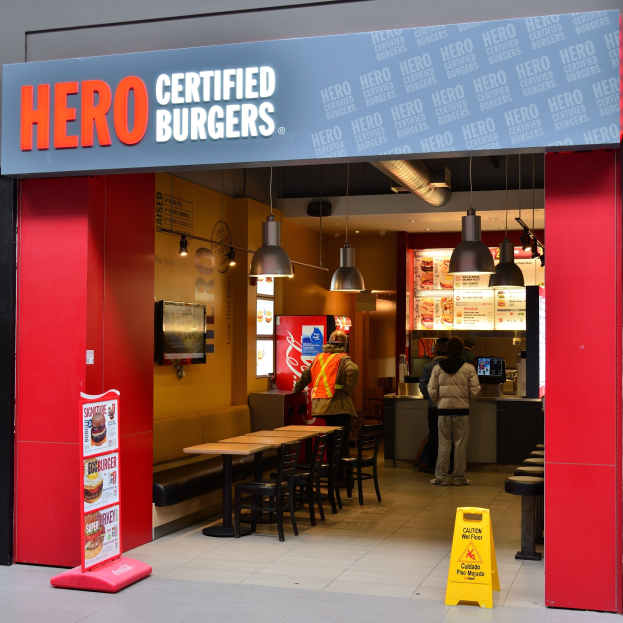Außenansicht eines Hero Certified Burgers Restaurants in London, England, mit Menschen drinnen, Tischen, Stühlen, einer Speisekarte, einem Wandfernseher und Deckenleuchten.
