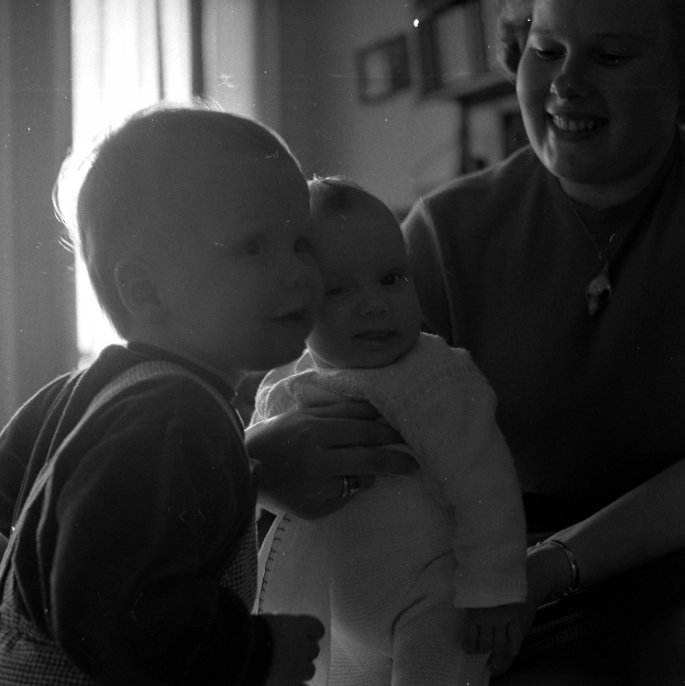 Eine Frau hält ein Baby mit einem warmen Lächeln, mit zwei anderen Kindern, die neben ihr stehen, in einem gemütlichen Raum mit Rahmen und einem Fenster im Hintergrund.