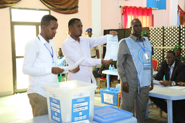 Eine Gruppe von Männern, die um einen Tisch mit verschiedenen Gegenständen stehen und bei einer Wahlurne abstimmen, mit Menschen und Möbeln im Hintergrund.