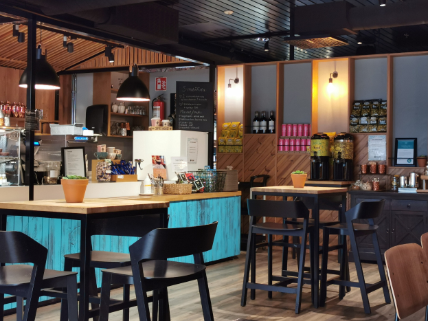 Innenraum eines Restaurants mit Holzmöbeln, einem Blumentopf auf einem Tisch, einer Schiefertafel, einem Kühlschrank, einem Feuerlöscher, Regalen mit Flaschen, Schränken mit Gegenständen, einem Rahmen mit einem Foto an der Wand und Deckenleuchten.