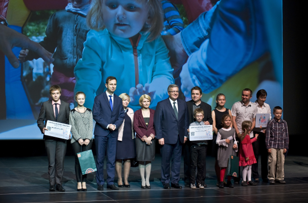 Gruppe von Menschen auf einer Buhne mit Taschen und Urkunden, im Hintergrund ein Bildschirm mit dem Bild eines Kindes und anderen.