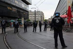 Polizeibeamte in schwarzen Uniformen und Helmen stehen entlang einer Straße, die von Glasfenster-Gebäuden, Bäumen, Polen, Drähten und einem klaren Himmel gesäumt ist, mit verstreuten Schirmen, Stühlen und Tischen.