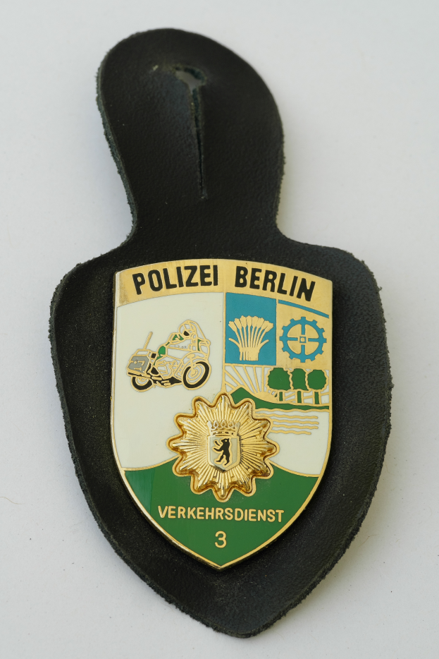 Ein Polizeistern mit der Aufschrift "Polizei Berlin" in fetter schwarzer Schrift auf einem blauen Schild mit einem weißen Stern und Rahmen, der auf einer Oberfläche liegt.