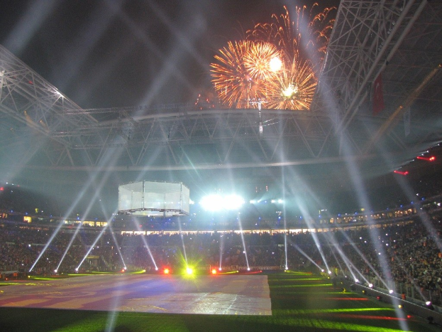 Hell erleuchtetes Olympisches Stadion in London bei Nacht mit grünem Rasen und einer großen Menge Menschen unter einem Feuerwerk-himmel.