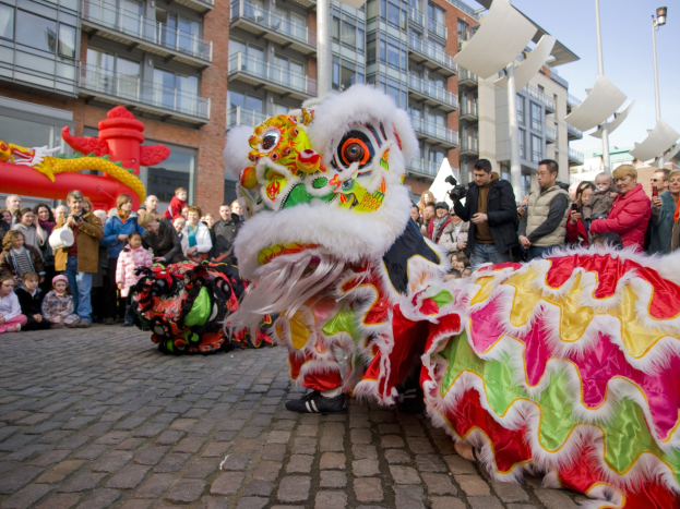 Ein lebendiges chinesisches Neujahrsfest in Amsterdam mit einer Löwen-Tanzvorstellung und einer Menge Schaulustiger, einige fotografieren das Ereignis, vor einer Kulisse aus Gebäuden, Laternenmasten und einem klaren blauen Himmel.