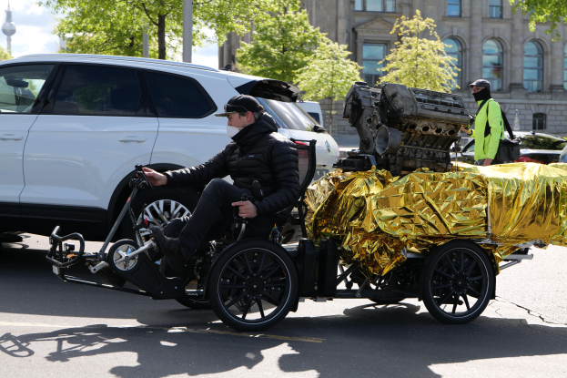 Ein Mann im Rollstuhl mit einem großen Motor daran, der eine schwarze Jacke und eine Mütze trägt, ist von Fahrzeugen auf einer Straße mit Bäumen, Gebäuden und Polen im Hintergrund unter einem klaren blauen Himmel umgeben.