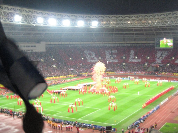 Ein Stadion mit Flutlicht, Eisengittern, Zuschauern, Personen am Boden, Werbetafeln, Anzeigetafeln, Überwachungskamera, Fahnen und Dekorationen.