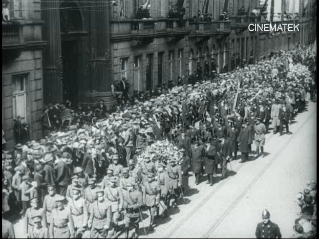 Schwarzes und weißes Foto einer großen Menge, die vor einem Gebäude die Straße entlangmarschiert, einige halten Gewehre in den Händen.