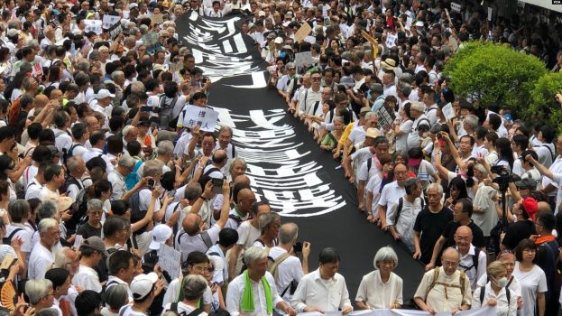 Eine große Gruppe von Menschen marschiert auf einer Straße, hält Protestschilder und Banner in der Hand, mit Topfpflanzen auf der rechten Seite und einem Gebäude im Hintergrund.