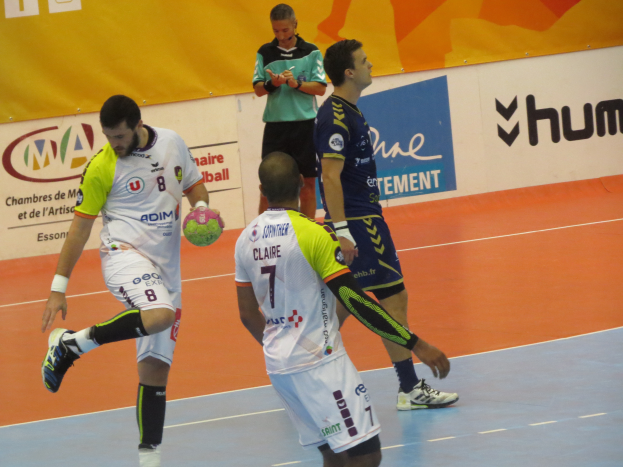 Eine Gruppe von Männern, die auf einem Handballfeld spielen, wobei einer einen Ball hält und im Hintergrund Schilder mit Text zu sehen sind.