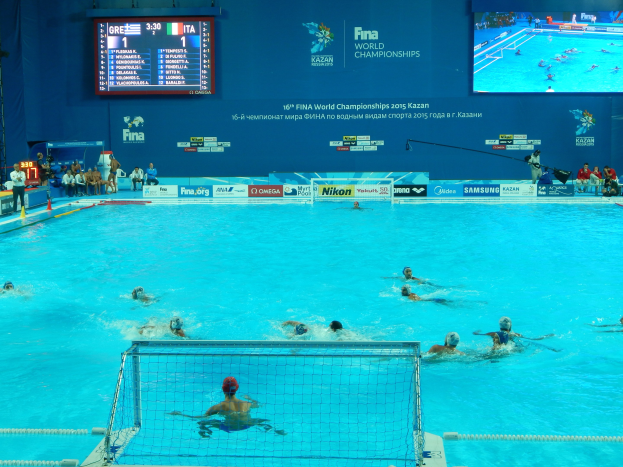 Gruppe von Athleten beim Wasserballspielen in einem Pool während der FINA-Weltmeisterschaften 2016 in Kazan, mit Zuschauern auf der linken Seite und einem Bildschirm mit Event-Branding im Hintergrund.
