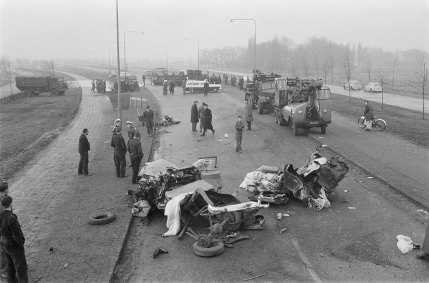 Schwarz-weiß-Szene eines Autounfalls am Straßenrand mit mehreren Fahrzeugen, Menschen drumherum, Laternenpfählen, Bäumen und sichtbarem Himmel.