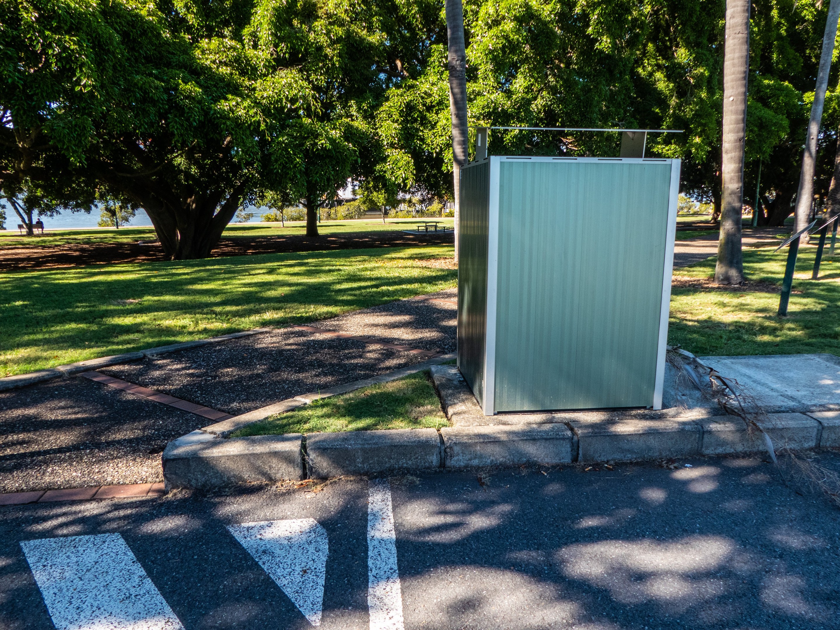 Transportable Toilette am Straßenrand in einem Park mit Bäumen, Gras, einem Gewässer und einem klaren blauen Himmel im Hintergrund.