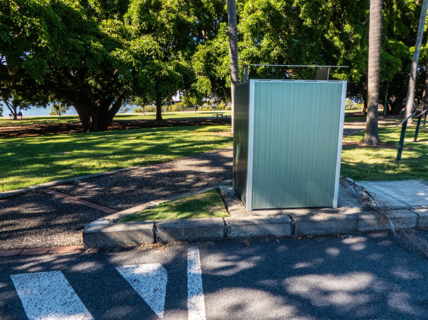 Transportable Toilette am Straßenrand in einem Park mit Bäumen, Gras, einem Gewässer und einem klaren blauen Himmel im Hintergrund.