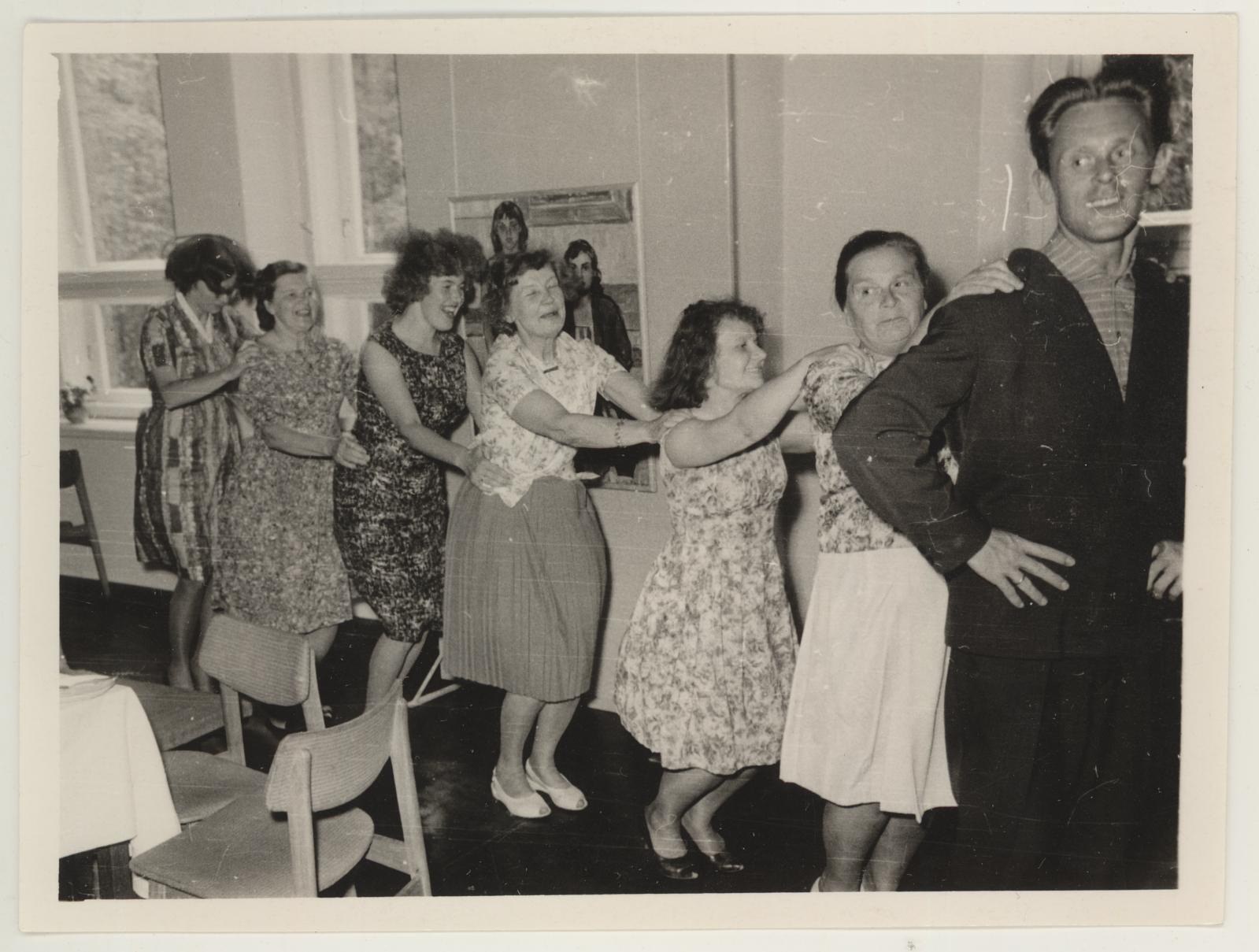 Eine Gruppe von Menschen, die in einem Raum im Stil der 1950er Jahre tanzen, mit Stühlen, einem Tisch mit wei├čer Decke, einem Foto auf der Wand und Fenstern im Hintergrund.