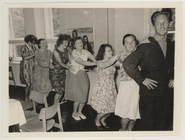 Eine Gruppe von Menschen, die in einem Raum im Stil der 1950er Jahre tanzen, mit Stühlen, einem Tisch mit wei├čer Decke, einem Foto auf der Wand und Fenstern im Hintergrund.