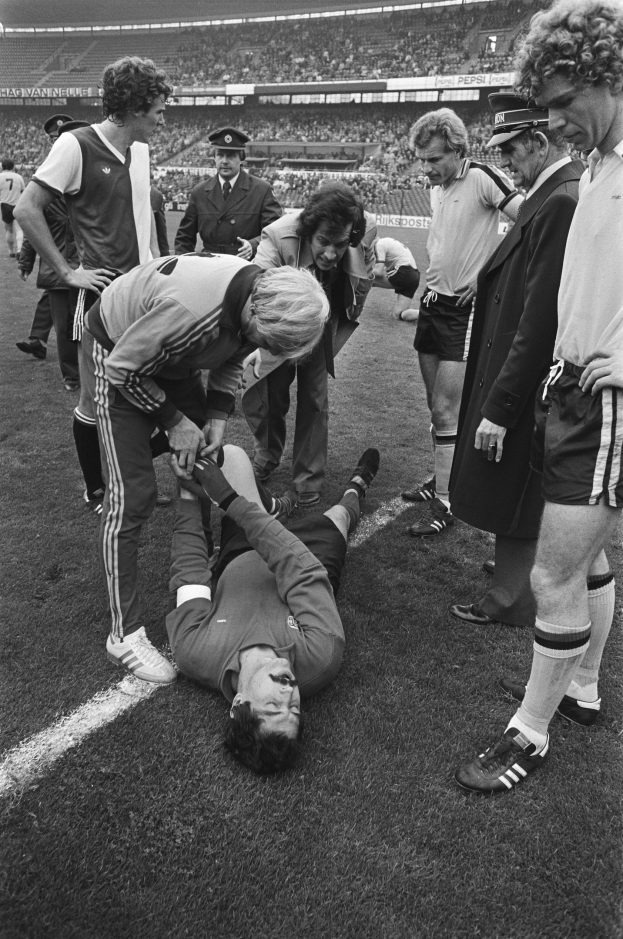 Schwarzes Foto eines Fußballspielers, der auf dem Boden liegt und von einer Gruppe Männer umgeben ist, mit einer Stadionkulisse im Hintergrund; der Spieler scheint verletzt zu sein.