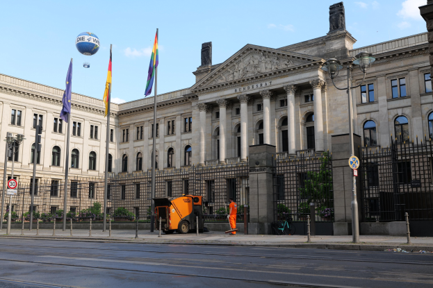 Großes modernes Gebäude mit vielen Fenstern, als Bundestag in Berlin identifiziert, umgeben von Straßeninfrastruktur, Fußgängern und Flaggen, unter einem bewölkten Himmel.