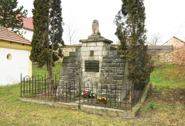 Ein steinerner Gedenkstein auf einem Rasenfeld, umgeben von einem Metallzaun und geschmückt mit Blumensträußen, mit Bäumen, Häusern und einer Wand im Hintergrund unter einem klaren blauen Himmel dient als Holocaust-Gedenkstätte in Sibiu, Rumänien.