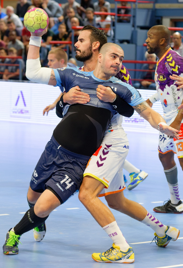 Männer spielen Hallenhandball in einer Arena, einer hält den Ball, während andere konkurrieren, mit Zuschauern auf Stühlen, Geländern und einer Wand im Hintergrund.