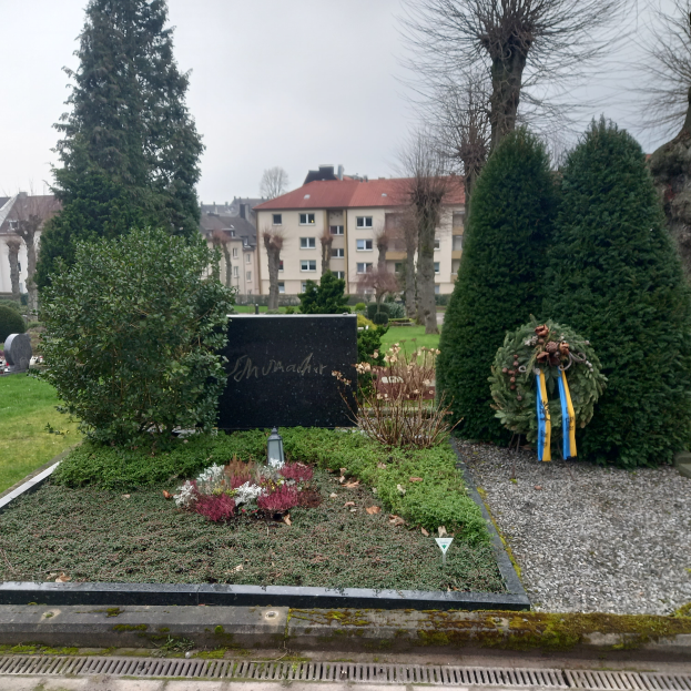 Ein Mahnmal für die Opfer des Zweiten Weltkriegs steht in einem Park, geschmückt mit einem Kränzchen und umgeben von Grün, mit Gebäuden im Hintergrund.