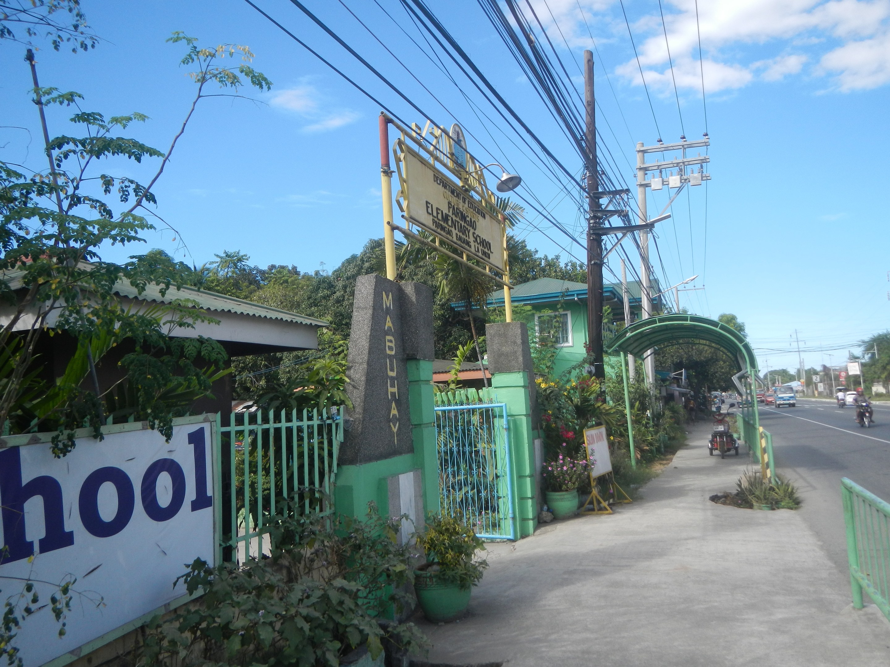 Einfahrt zu einer Schule mit einem "Schule"-Schild, Fahrzeuge auf der Straße, ein Torbogen, ein Tor, eine Namensplakette, Pflanzen, Bäume, Strommasten mit Drähten und eine bewölkte Himmel im Hintergrund.