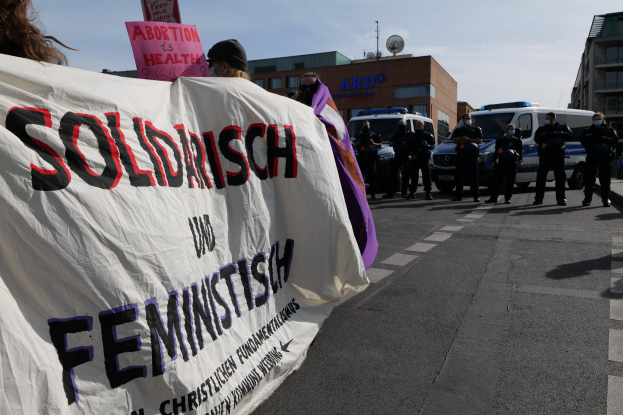 Gruppe von Menschen, die eine 'Solidarität und Feminismus'-Schlagzeile tragen, mit geparkten Fahrzeugen, Gebäuden, einer Schüsselantenne und einem klaren blauen Himmel im Hintergrund.