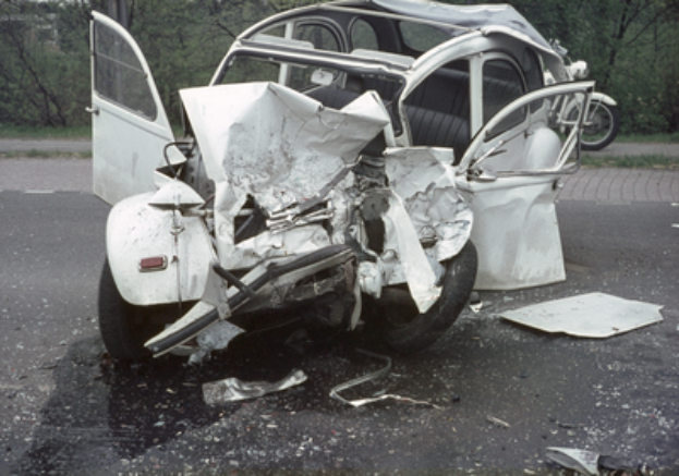 Ein verunglücktes Auto mit schwerer Frontschaden steht am Straßenrand, mit Bäumen, einem Gebäude und einem anderen Fahrzeug im Hintergrund.