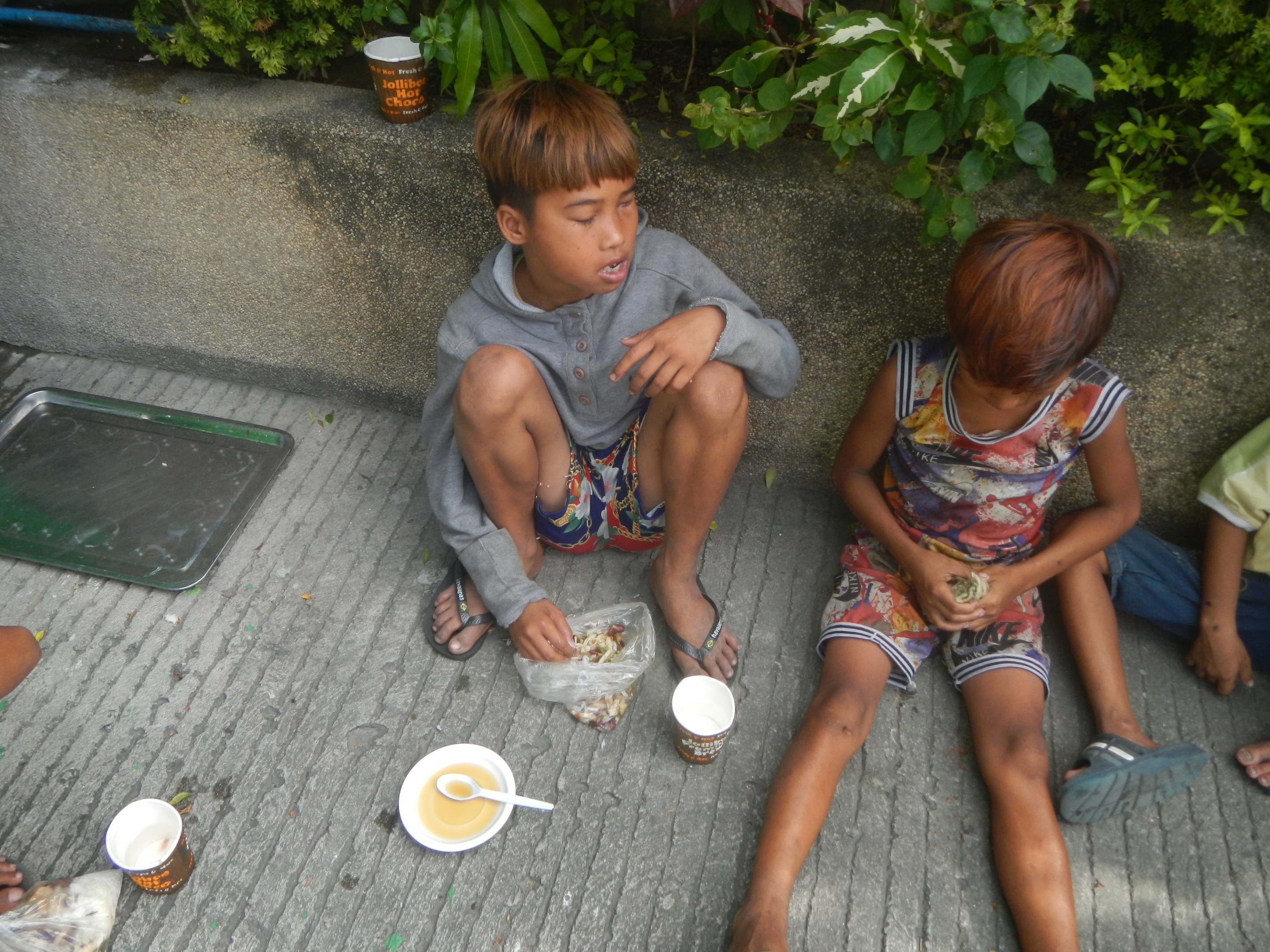 Gruppe von Kindern, die auf dem Boden sitzen und aus Schüsseln mit Löffeln essen, mit einem Jungen in der Mitte, umgeben von anderen, Pflanzen und einer Wand im Hintergrund und einem Tablett auf der linken Seite.