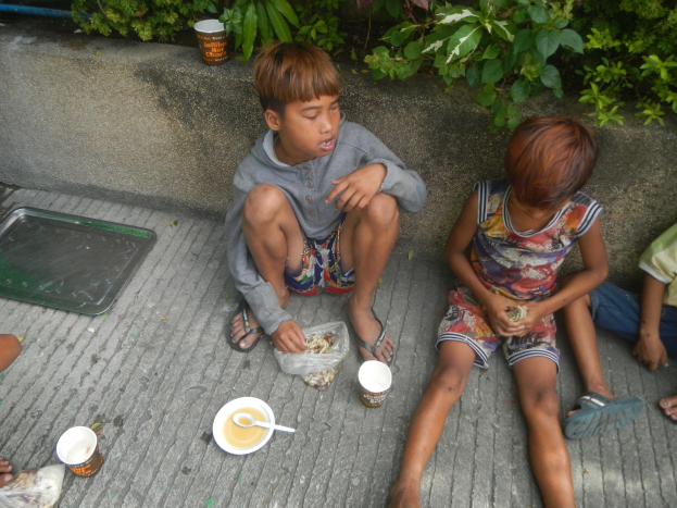 Gruppe von Kindern, die auf dem Boden sitzen und aus Schüsseln mit Löffeln essen, mit einem Jungen in der Mitte, umgeben von anderen, Pflanzen und einer Wand im Hintergrund und einem Tablett auf der linken Seite.