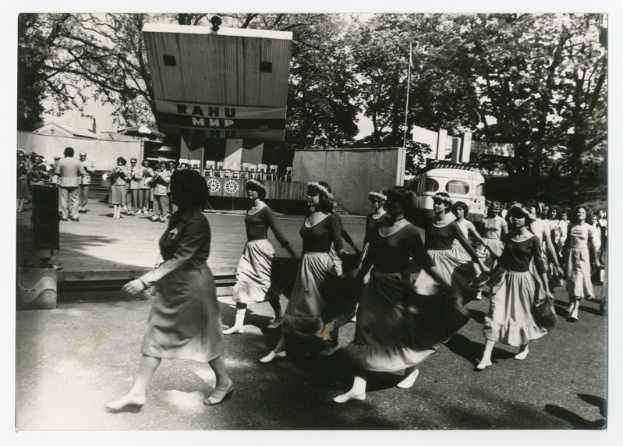 Eine Gruppe von Frauen, die auf der Straße tanzen, umgeben von einer Menge, einem Lautsprecher, einem Fahrzeug, einer Tafel mit Text, einem Fahnenmast, Gebäuden und einem bewölkten Himmel.
