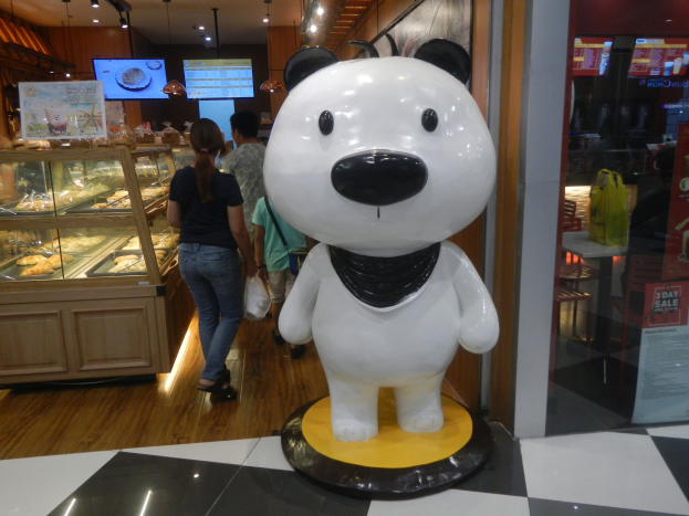 Eine Riesengiant-Panda-Statue steht in einem Laden umgeben von Einkaufenden, mit einer Auslage mit Lebensmitteln links und an der Decke montierten Fernsehern und Lichtern im Hintergrund.