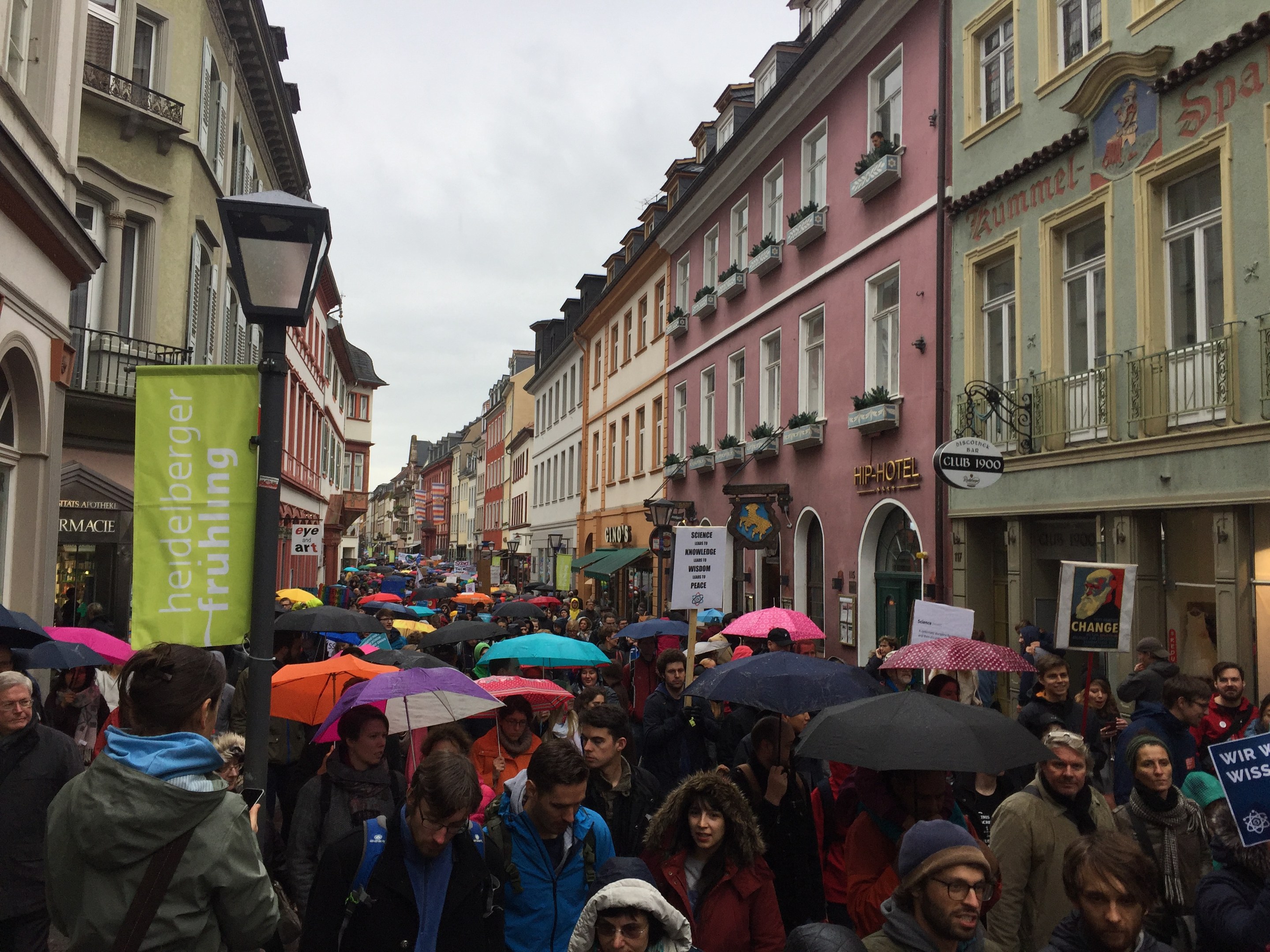 Große Gruppe von Menschen geht mit Schirmen auf der Straße, einige tragen Taschen und halten Schilder, mit Gebäuden und einem Lichtmast im Hintergrund.