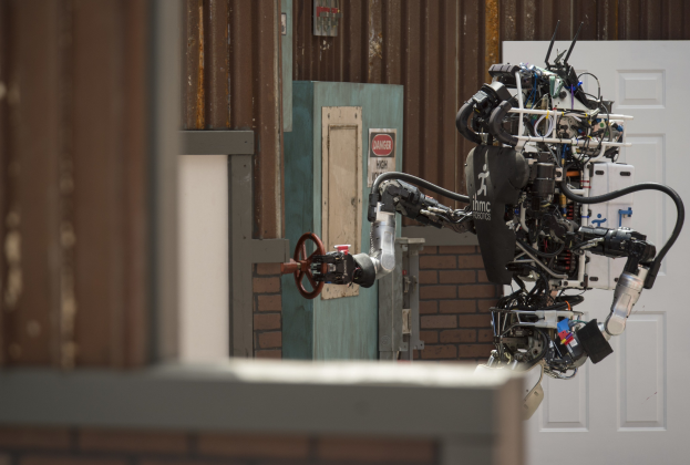 Humanoider Roboter mit ausgestreckten Armen und geneigtem Kopf vor einem Gebäude mit einer Tür und einer Wand im Hintergrund