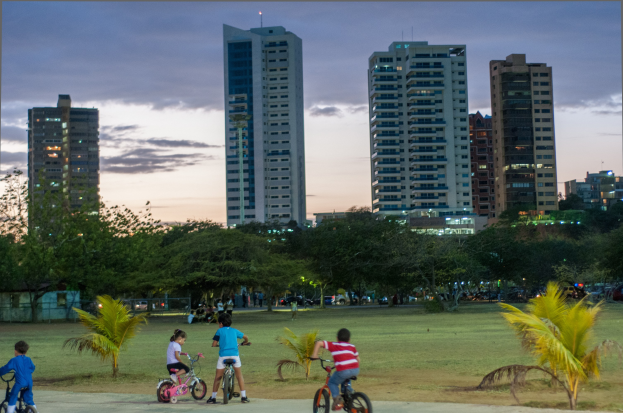 Kinder auf Fahrrädern in einem Park bei Sonnenuntergang, mit Hochhaussilhouetten vor einem farbenfrohen Himmel