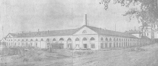 Schwarz-weiß-Foto eines großen, mehrfenstrigen Gebäudes, das von Bäumen und Pflanzen umgeben ist und unter einem klaren Himmel steht, mit Text unten, der "1892 – das erste Fabrikgebäude in der Stadt" lautet.