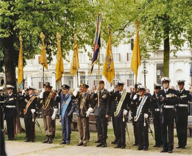 Gruppe uniformierter Männer in Formation während einer Regimentszeremonie, einige halten Gewehre, mit Fahnen, Bäumen und Laternenmasten im Hintergrund.