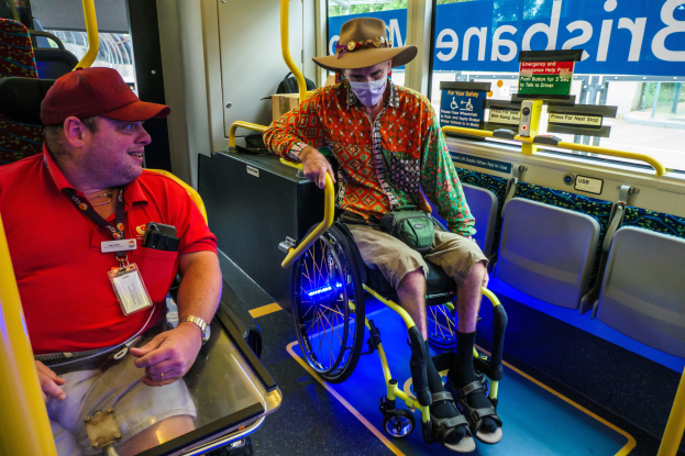 Zwei Männer in Rollstühlen auf einem Bus, einer trägt ein rotes T-Shirt und eine Kappe, der andere ein orangefarbenes Hemd und eine braune Mütze, beide tragen Masken und Ausweise, mit gelben Stangen und Glasfenstern, die Bäume, Gebäude und Himmel zeigen.