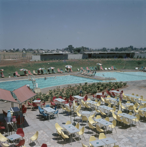 Ein Freibad mit Tischen, Stühlen und Sonnenschirmen, umgeben von Blumenpflanzen, einer Mauer, einer Wand, einer Baumgruppe, einigen Gebäuden und einem bewölkten Himmel.