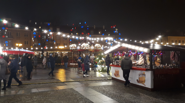 Ein belebter Weihnachtsmarkt bei Nacht in einer Stadt mit Menschen, geschmückten Ständen, Gebäuden, Bäumen und festlich beleuchtetem Himmel.