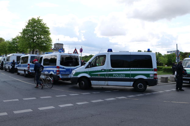 Polizeifahrzeuge stehen entlang einer Straße mit einem Radfahrer im Vordergrund, Bäume, Strommasten, Schilder, Gebäude und ein bewölkter Himmel im Hintergrund.