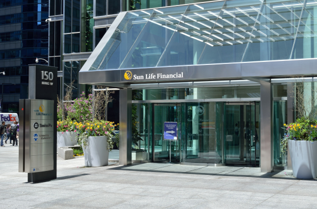 Moderner Glasbau der Sun Life Financial in Toronto mit einer Leuchttafel, Topfpflanzen mit Blumen, Fußgängern, einer Straßenlaterne, einer Leuchttafel, Fahrzeugen auf der Straße und einem bewölkten Himmel.