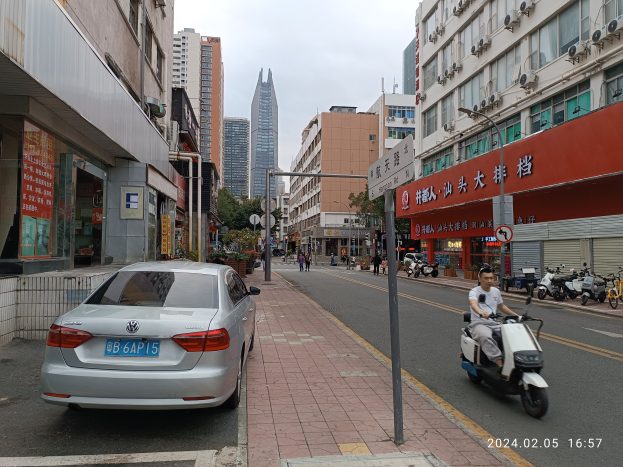 Ein Mann fährt mit einem Roller eine Straße mit hohen Gebäuden entlang, mit Menschen auf den Gehwegen und Fahrzeugen auf der Straße, unter einem klaren blauen Himmel mit Bäumen im Hintergrund.