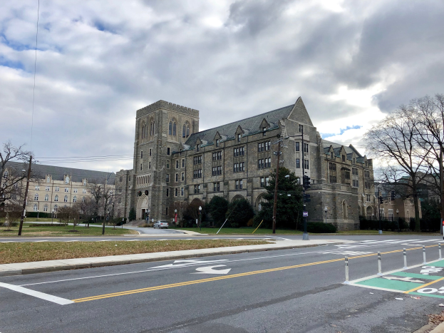 Großes Gebäude mit vielen Fenstern, als Universität von Pennsylvania beschriftet, umgeben von Bäumen, Straßenlaternen, Verkehrsschildern und Fahrzeugen an einem bewölkten Tag.