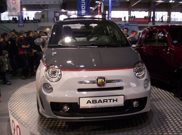 Ein roter Fiat 500 Abarth auf einer Autauschstellung mit Menschen drumherum und Bannern im Hintergrund.